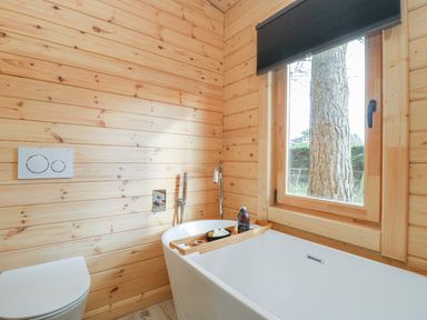 A bathroom with a white bathtub a wooden bath tray a bottle a white toilet and a window showing a tree at Gardeners Lodge in Huby