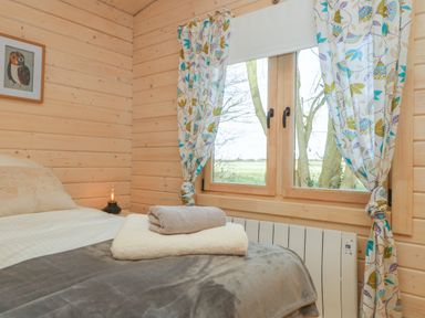 A bedroom with a double bed gray blanket folded towels on bed a window with patterned curtains and a framed owl picture on wood paneled walls at Gardener's Lodge in Huby