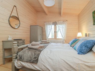 A bedroom with a double bed, pillows, folded towels, a window with curtains, a side table with a lamp, a dresser, a wall mirror, and wooden walls at Gardeners Lodge in Huby