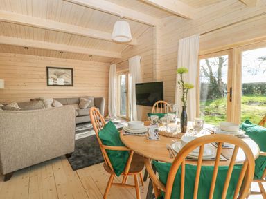 A dining table with green cushions on chairs and a vase with flowers beside a living area with a sofa and a wall mounted tv at Gardener's Lodge in Huby