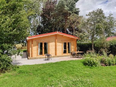 A wooden cabin with two sets of glass doors in a garden with grass trees and outdoor furniture at Gardeners Lodge in Huby