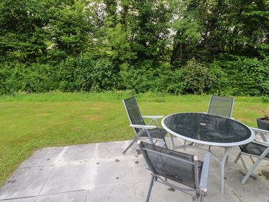 An outdoor patio area with a round glass table and four black mesh chairs beside a grassy lawn and trees at 11 Tremore in Liskeard