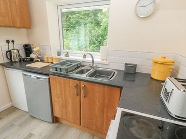 A kitchen with a sink, countertop appliances, wooden cabinets, and a window overlooking greenery at 11 Tremore in Liskeard