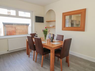 A dining area with a wooden table and six brown chairs with placemats a vase of flowers a bottle and two glasses a wooden framed mirror on the wall and a window showing an outside wooden structure at Hazonleigh in Seahouses