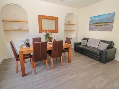 A dining area with a wooden table and six chairs next to a black sofa with cushions and a picture on the wall at Hazonleigh in Seahouses