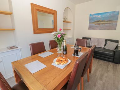 A dining area with a wooden table set with croissants flowers a bottle and glasses and a black sofa with cushions and a blanket at Hazonleigh in Seahouses