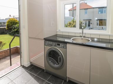 A kitchen area with a washing machine under a counter with a sink and window and an open door showing a garden at Hazonleigh in Seahouses