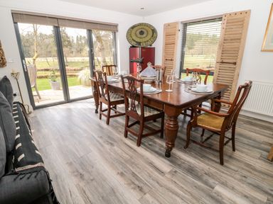 A dining room with a wooden table and six chairs next to sliding glass doors and a window with wooden shutters at Romsey Lodge in Wark