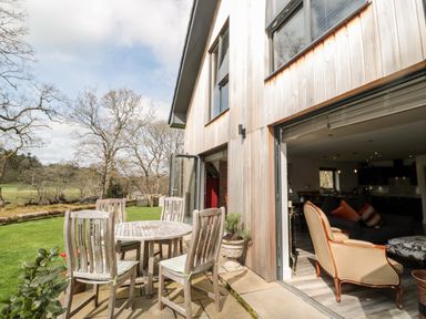 An outdoor patio with wooden chairs and table next to a modern wooden house with open doors showing a living area at Romsey Lodge in Wark