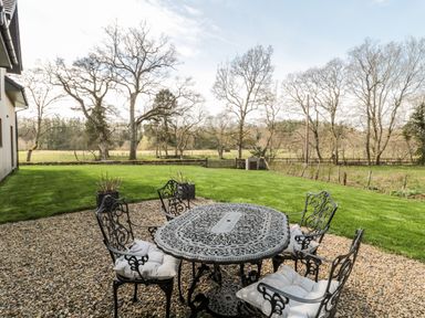 An outdoor patio area with a metal table and four chairs with cushions on a gravel surface next to a lawn and trees at Romsey Lodge in Wark