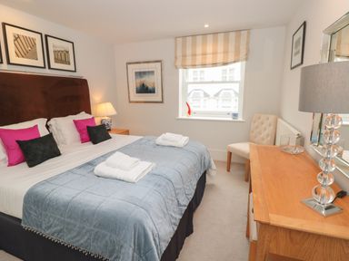 A bedroom with a double bed with pillows, a wooden dresser, a chair, a window with blinds, and framed pictures on the walls at 42 Dart Marina Dartmouth