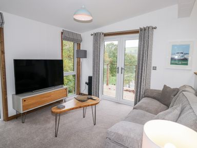 A living room with a large TV on a stand a coffee table sofa and glass doors with curtains at 1 Valley View in Lanreath