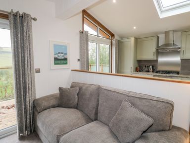 A grey sofa with cushions near a half wall and glass doors with curtains in a kitchen living area at 1 Valley View in Lanreath