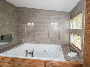 A bathroom with a large jacuzzi tub surrounded by tiled walls and two windows with blinds at 1 Valley View in Lanreath