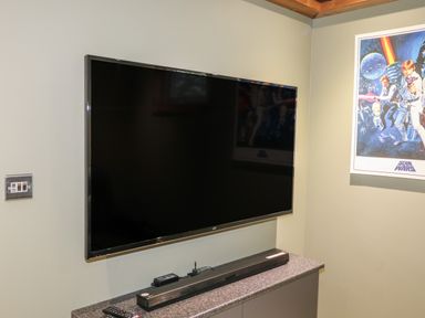 A wall-mounted television above a cabinet with a soundbar remote and power adapter next to a Star Wars poster at 1 Valley View in Lanreath
