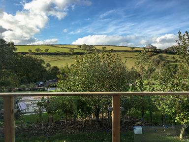 A view of green fields and trees seen from behind a wooden balcony railing at 1 Valley View in Lanreath