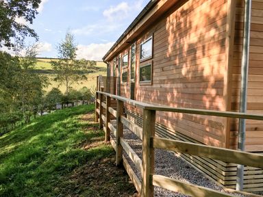 An exterior view of a wooden house with a fenced path and grassy hillside in the background at 1 Valley View in Lanreath