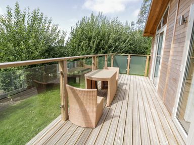 An outdoor balcony with wooden flooring a glass and wood railing and a wicker table with two chairs at 1 Valley View in Lanreath