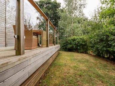 A wooden deck with outdoor wicker chairs and a glass railing next to a grassy area with trees and bushes at 1 Valley View in Lanreath