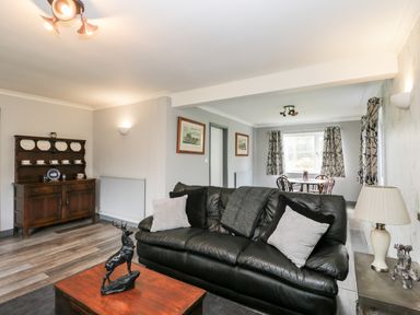 A living room with a black leather sofa and cushions a wooden coffee table with a deer statue a side table with a lamp and sculpture a wooden cabinet with china plates and cups a dining table with chairs near windows with curtains at Pool House in The Narth near Trellech
