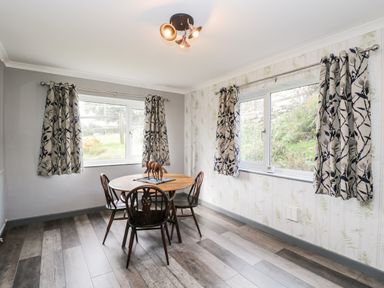 A dining room with a round wooden table and four chairs near two windows with patterned curtains at Pool House in The Narth near Trellech