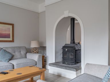 A living room with a wood burning stove a gray sofa a wooden coffee table and a table lamp at Keepers Cottage Hawkshead