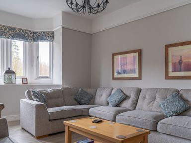 A living room with a grey sectional sofa blue patterned cushions wooden coffee table two framed pictures and a window with patterned valance at Keepers Cottage in Hawkshead