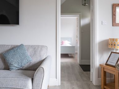 A gray sofa with a blue patterned pillow in a room with a wooden side table lamp and a view into a bedroom with a gray bed at Keepers Cottage in Hawkshead