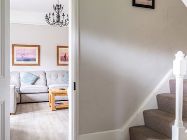 A view through an open door showing a living room with a sofa cushions wooden coffee table wall art and a chandelier next to a carpeted staircase at Keepers Cottage Hawkshead