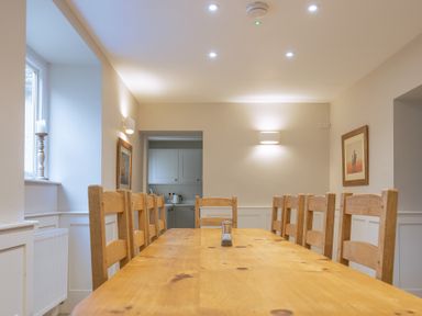 A dining room with a long wooden table and chairs near a window with candle and a kitchen in the background at Keepers Cottage in Hawkshead