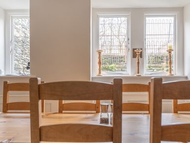A dining area with wooden chairs and a table by three windows with candlesticks at Keepers Cottage in Hawkshead