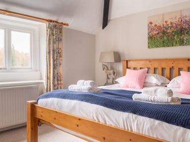 A bedroom with a wooden bed with pink pillows white towels and a blue blanket next to a window with floral curtains at Keepers Cottage Hawkshead
