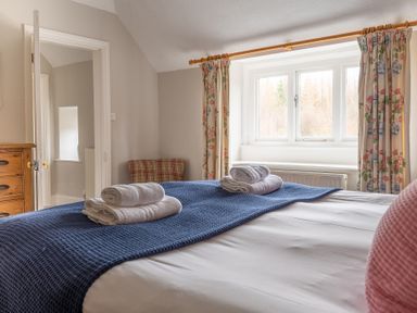 A bedroom with a made bed with towels on a blue runner a wooden dresser a window with floral curtains and a plaid chair at Keepers Cottage in Hawkshead