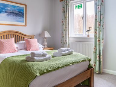 A bedroom with a wooden bed green blanket pink pillows bedside lamp and window with floral curtains at Keepers Cottage in Hawkshead