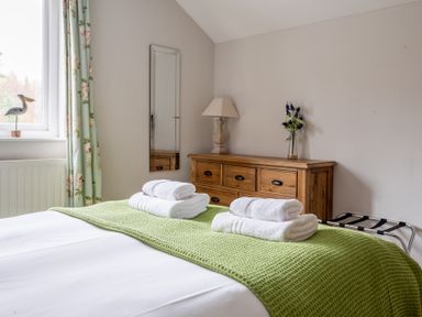 A bedroom with a bed covered with a green blanket and white towels on top a wooden dresser with a lamp and flowers and a window with floral curtains at Keepers Cottage in Hawkshead