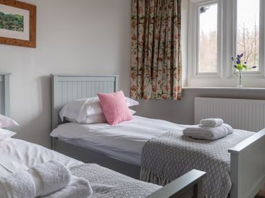 A bedroom with two single beds with white linens and pink and white pillows a window with floral curtains and a vase of flowers on the windowsill at Keepers Cottage in Hawkshead