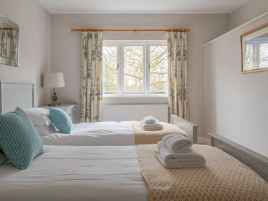 A bedroom with two single beds two blue pillows folded towels a lamp and windows with floral curtains at Keepers Cottage in Hawkshead