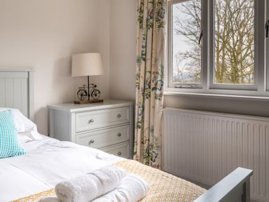 A bedroom with a bed white linens and towels bedside table with lamp and window with floral curtains at Keepers Cottage in Hawkshead