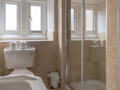 A bathroom with a toilet, shower with glass door, two windows, and a small trash bin at Keepers Cottage in Hawkshead