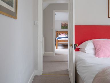 A bedroom with a red headboard and pink pillow visible through a doorway showing another bedroom with a wooden bed frame and bedding at Keepers Cottage in Hawkshead
