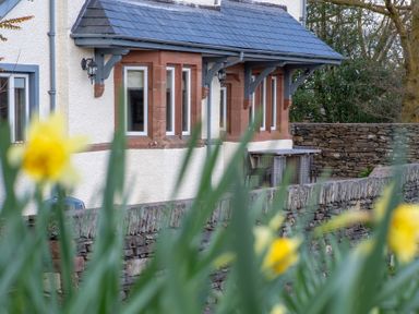 A stone wall with yellow flowers in front of a house with multiple windows and outdoor seating at Keepers Cottage in Hawkshead