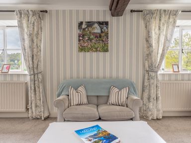 A living room with a beige sofa and striped cushions between two windows with floral curtains and a coffee table with a book at Grassholme in Hawkshead