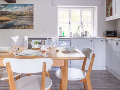 A dining area with a wooden table set for four with plates and glasses in a kitchen with white cabinets and a window at Grassholme in Hawkshead