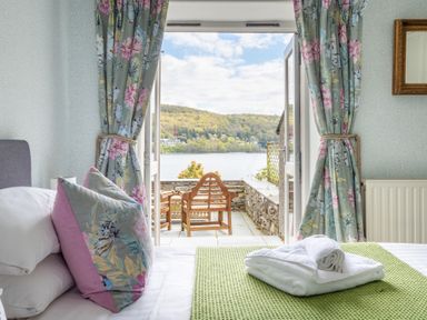 A bedroom with a bed and floral curtains opening to a terrace with wooden chairs overlooking a lake at Grassholme in Hawkshead