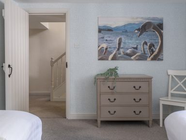 A room with a taupe chest of drawers with a plant on top a white chair an open door showing a staircase and a painting of swans on the wall at Grassholme in Hawkshead