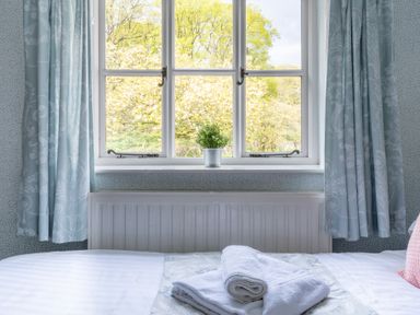 A bedroom window with blue patterned curtains and a plant on the windowsill above a bed with folded white towels at Grassholme in Hawkshead