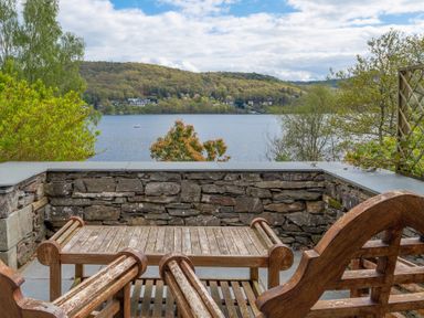 A stone patio with wooden chairs and a table overlooking a lake and wooded hills at Grassholme in Hawkshead