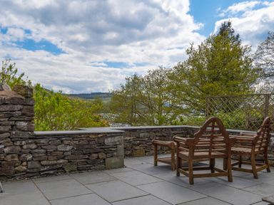 An outdoor stone patio with wooden bench seating and stone wall surrounded by trees and hills at Grassholme in Hawkshead