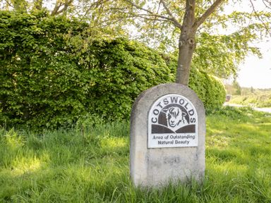 A stone sign with a sheep illustration reading Cotswolds Area of Outstanding Natural Beauty on grass near a tree and hedge at Rosemead in Moreton-In-Marsh