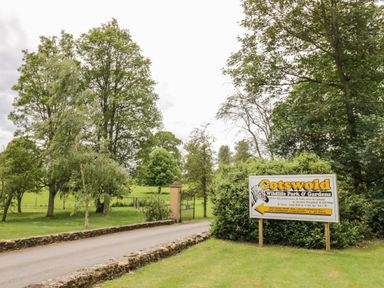 A road with stone walls on each side leading to a gated entrance surrounded by trees and a sign for Cotswold Wildlife Park and Gardens in Moreton-In-Marsh